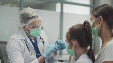 Tecrübeli bir çocuk doktoru küçük bir kız için rutin bir aşı yapar, şırıngadan hava salıp omzuna antiseptik koyar, anne kızını kucağında tutar.