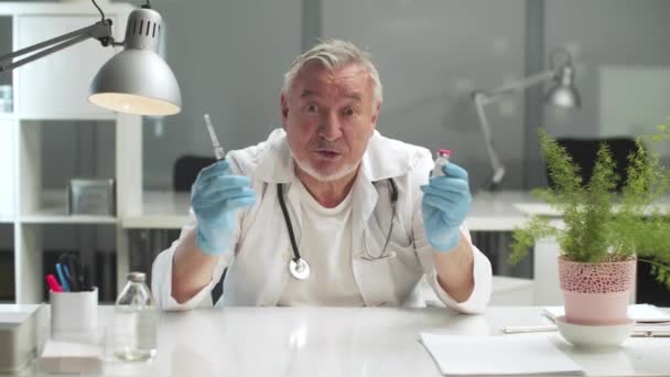 Un medic cu părul gri din clinică vorbește despre vaccinare și arată vaccinul și seringa. — Videoclip de stoc