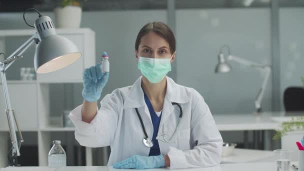 Un medic de sex feminin dintr-o clinică stă la o masă și arată o sticlă de vaccin camerei. — Videoclip de stoc