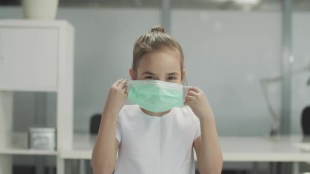 A little girl in a white T-shirt puts on a medical mask and adjusts it on her face — Stock Video