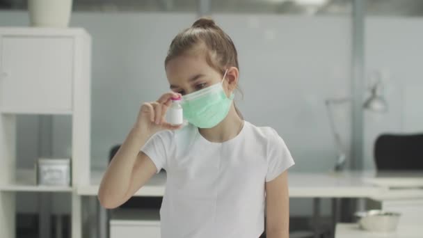 Nello studio medico, una bambina con una maschera medica esamina una bottiglia di vaccino. — Video Stock