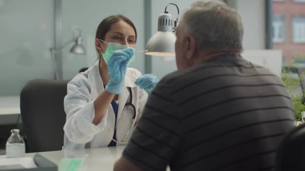 Χαριτωμένο κορίτσι βοηθός εργαστηρίου στην κλινική παίρνει ένα δείγμα από το στόμα ενός ηλικιωμένου ασθενή για ανάλυση σε covid-19 — Αρχείο Βίντεο