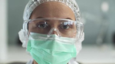 Genç bir kadın doktorun yakın plan portresi ve tıbbi maske, şapka ve gözlük.