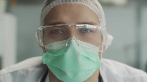 Ritratto emotivo ravvicinato di un medico mascherato che combatte la diffusione di un'infezione coronavirica. Occhi azzurri e occhiali protettivi di plastica, — Video Stock