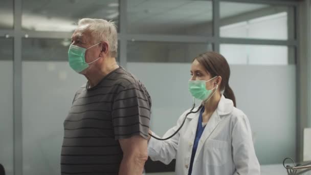 Menina terapeuta realiza ausculta dos pulmões a partir das costas de um paciente idoso com pneumonia — Vídeo de Stock