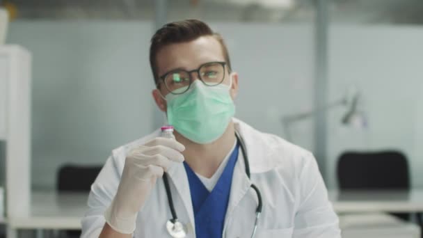 De arts beveelt de patiënt een nieuw coronavirusvaccin aan. Een blik op de camera. Een fles medicijnen in zijn hand. Latex handschoenen en masker, slow motion. — Stockvideo