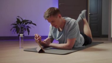 Bir adam yoga minderinde yatar ve tablet üzerinde egzersiz yapar.