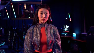 Parlak ceketli katı bir Asyalı kadın neon ışıklı bir odada kameraya bakar.