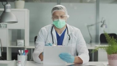 Koruyucu takım elbiseli, tıbbi şapkalı ve eldivenli genç bir doktorun yakın plan portresi bir devlet hastanesinin ofisinde kalemle not defterine not alır. Bulanık arkaplan, yavaş çekim.