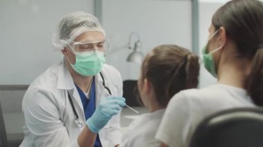 Genç bir erkek laboratuvar asistanı, covid-19 analizi için küçük bir kızın nazofarnx 'inden örnek alır. Annesi tarafından desteklenir.