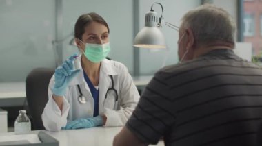 Genç bir kadın doktor yaşlı hastasına grip ve koronavirüse karşı aşı yapılmasını öneriyor. Elinde bir şişe aşı tutuyor.