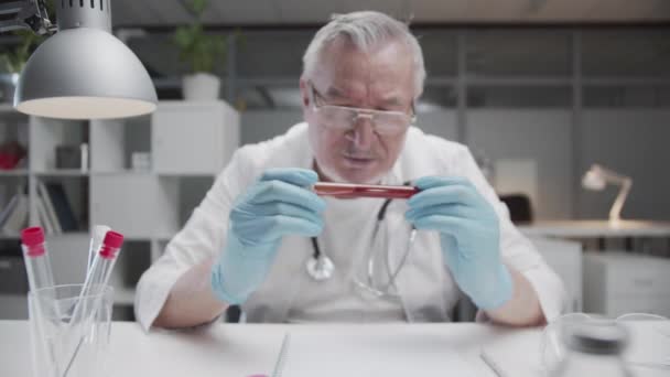 Ein alter Arzt in der Arztpraxis hält ein Reagenzglas mit einem Bluttest eines Patienten mit Coronavirus in der Hand. Prüft den Bluttest, um eine Behandlung zu verschreiben. — Stockvideo