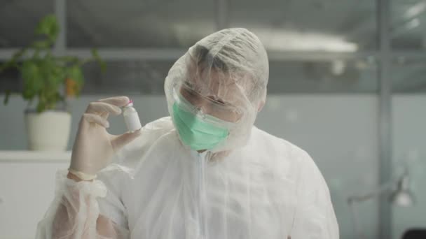 Un médico con traje de protección médica lleva una botella de vacuna contra el coronavirus en la mano y la examina. — Vídeos de Stock