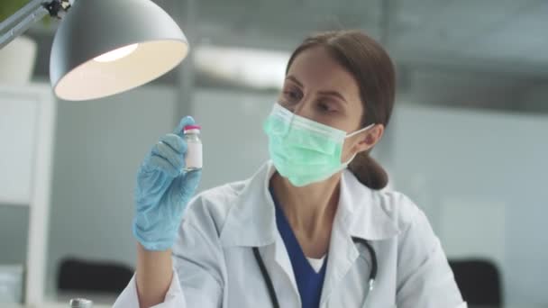 A pretty masked medical girl examines a bottle of vaccine under a lamp — Stock Video