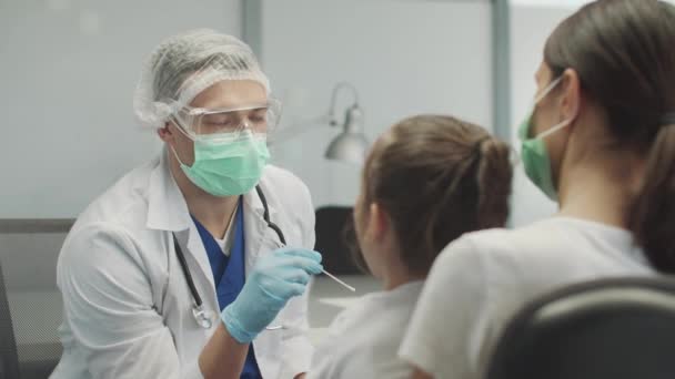 Un joven ayudante de laboratorio toma un hisopo de la nasofaringe de una niña para su análisis en covid-19, ella es apoyada por su madre — Vídeos de Stock