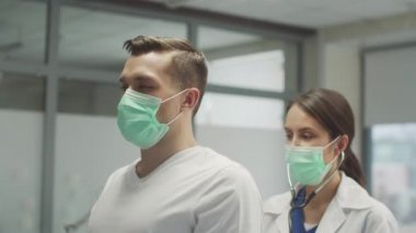 Maskeli genç bir adam öksürük şikayetiyle doktora gitti. Kadın bir doktor ciğerlerini arkadan dinliyor.