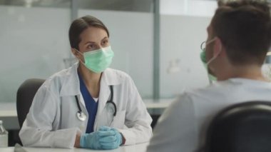 Maskeli ve eldivenli genç bir bayan doktor bir hastayı muayene ediyor. Genç adama virüse karşı aşı ihtiyacını anlatıyor..