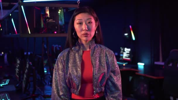 A strict young asian woman in a shiny jacket looks at the camera in a room with a neon light — Stock Video
