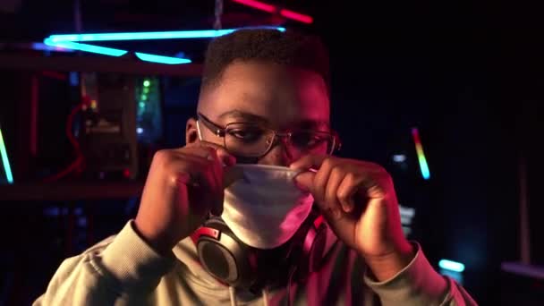 Ung svart kille bär glasögon på dator klubb sätter på medicinsk mask för skydd under pandemi — Stockvideo