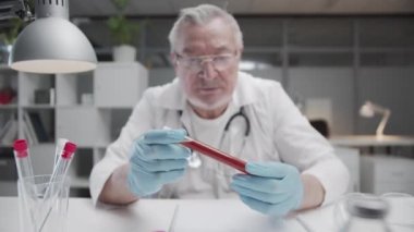 Klinikteki deneyimli bir doktorun elinde bulunan bir test tüpündeki kan örneğinin analizi. Doktor test sonuçlarını cam şişede inceliyor, yavaş çekimde.