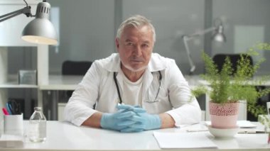 Yaşlı bir tıp profesörü masasında oturuyor, eldivenli elleri masanın üzerinde dinleniyor.