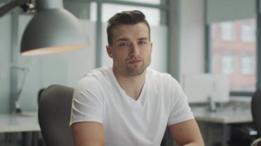 Ciddi bir genç erkek girişimci ya da serbest çalışan ofiste kameraya bakar. İş yerinin iç kısmında yakın plan portre.