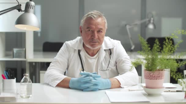 연로 한 의학 교수가 책상에 앉아 장갑낀 손을 책상 위에 놓고 앉아 있다 — 비디오
