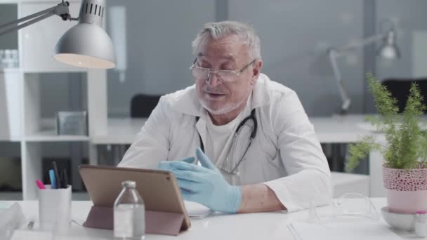 Un médico de cabellos grises y confiable consulta a un paciente en línea a través de una videollamada en una tableta sobre medidas de protección antiviral. Máscaras médicas y guantes de látex. — Vídeos de Stock