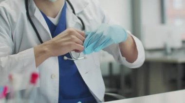 Yüzü olmadan yakın çekim. Boynunda steteskop olan bir doktor hastane ofisinde masasında lateks eldiven çıkartıyor..