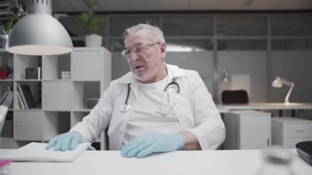 疲れた高齢者の経験豊富な医師は、医務室の職場で彼の一日を終え、彼の眼鏡を脱いでいます。コロナウイルスとの戦いと患者の受信. — ストック動画