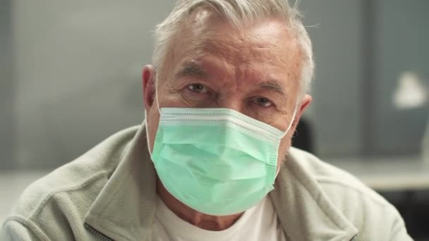 Siwy mężczyzna w masce medycznej patrzy w kamerę, portret z bliska — Wideo stockowe