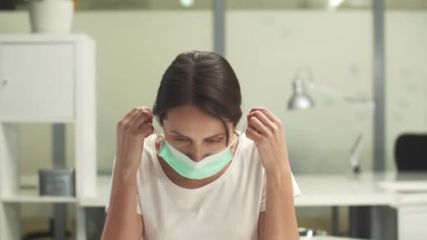 Une jeune femme s'assoit à un bureau dans le bureau, enlève son masque médical et sourit à la caméra — Video