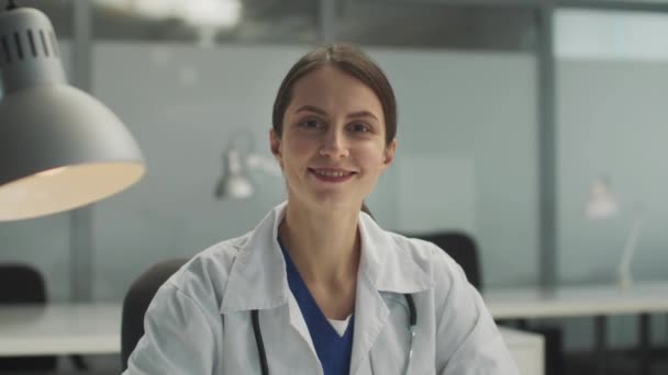 Девушка-интерн в больнице работает в первый день, позирует перед камерой за столом, улыбается — стоковое видео