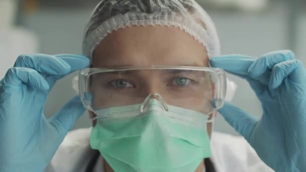 Närbild porträtt av en smittsam sjukdom läkare i en morgonrock, medicinsk mask och handskar. Läkaren tar av sig skyddsglasögonen och tittar på kameran med ett trött utseende. — Stockvideo
