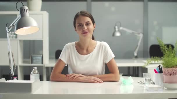 办公室里漂亮的年轻女人，穿着一件白色的T恤衫，坐在办公桌前，看着摄像机 — 图库视频影像