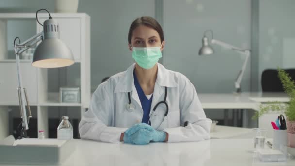 Příjemná doktorka v lékařské masce a rukavicích sedí za stolem v kanceláři, ruce v rukavicích složené na stole — Stock video