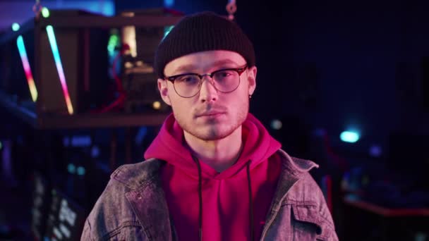 Een jonge gamer in een hoodie, denim jas, gebreide hoed en bril staart aandachtig naar de camera — Stockvideo