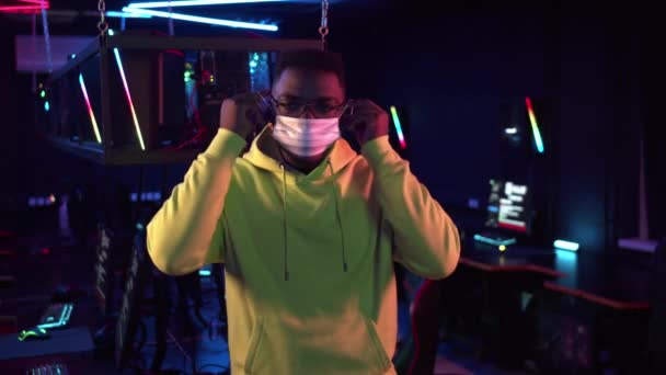 In de computerruimte tijdens de pandemie, ter bescherming van de spelers, is het gebruikelijk om medische maskers te dragen, de zwarte man zet een masker op zijn gezicht — Stockvideo