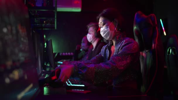 Молодые люди в медицинских масках пришли в компьютерный клуб во время пандемии коронавируса, девушка садится за компьютер, надевает наушники и выбирает игру — стоковое видео