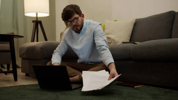 Un hombre europeo está escribiendo en un portátil y mirando los papeles sentados en el suelo. — Foto de Stock