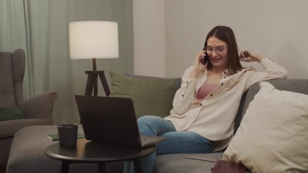 Женщина в перерыве разговаривает по телефону, сидя на диване в гостиной. — стоковое видео