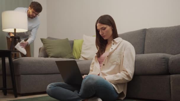 Мужчина убирает комнату, пока девушка работает за компьютером.. — стоковое видео