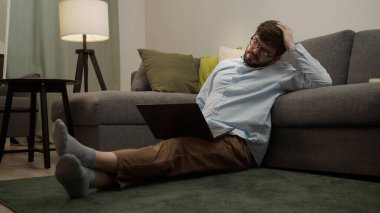 Yorgun bir adam başını tutup dizüstü bilgisayarla yere oturur..