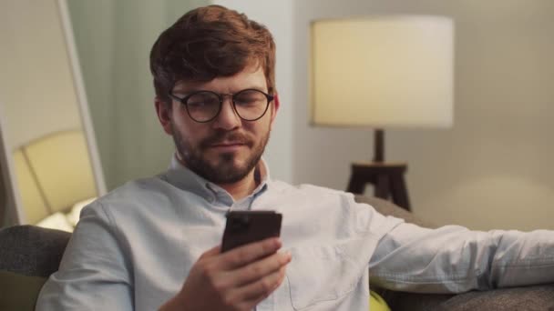 Homem europeu olha para o telefone enquanto sentado no sofá. — Vídeo de Stock