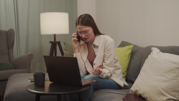 Wanita Eropa berbicara di telepon sambil duduk di sofa dan melihat laptopnya — Stok Video