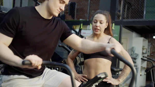 A strong sportsman is exercising on an exercycle and an athletic woman is talking with him