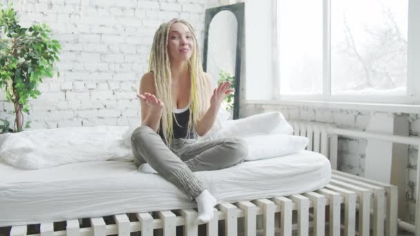 Een vrouw met dreadlocks en huiskleding zit op een groot bed en praat met de camera — Stockvideo