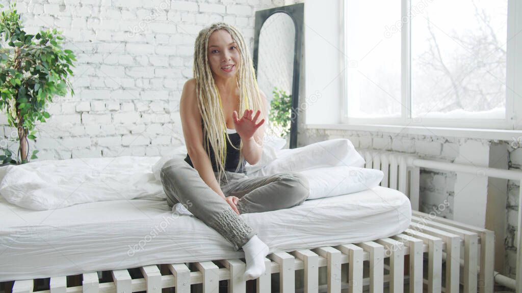 A woman with dreadlocks and home clothes sits on a large bed and talks to the camera