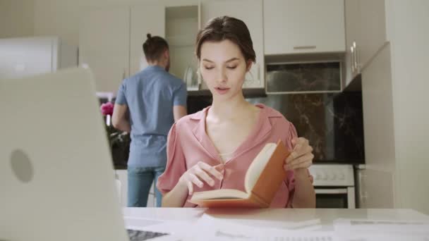 Egy felnőtt nő dolgozik és böngészi a könyvet, miközben a férje házimunkát végez a háttérben. — Stock videók