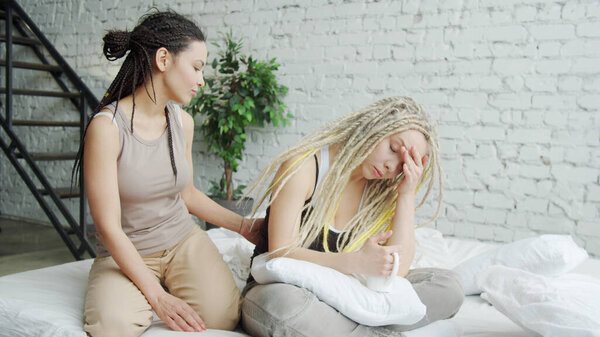 Depressed girl is sitting in bed while her girlfriend is trying to calm her down